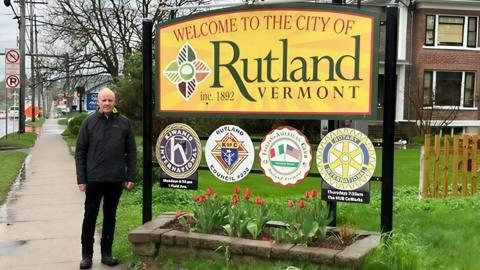 An image of a man in dark trousers and a dark coat standing by a sign for Rutland in Vermont.