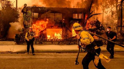 A firefighter drags a hose towards a burning house in the Altadena area of Los Angeles, while another firefighter looks on