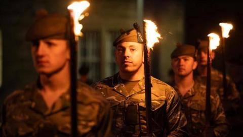 A line of four solemn soldiers holding lit torches under cover of darkness 