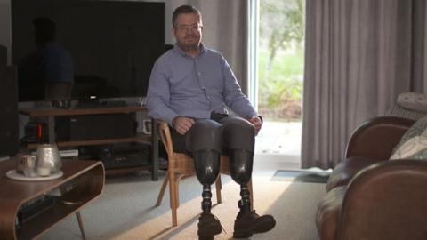 Neil Hopper is sitting in his lounge. He has prosthetic legs and is wearing a blue shirt and a pair of glasses.