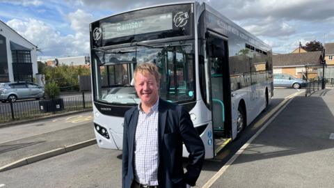A man wearing a navy jacket and a checked shirt stands in front of a white bus which says 'Ramsey' on it's destination board. 