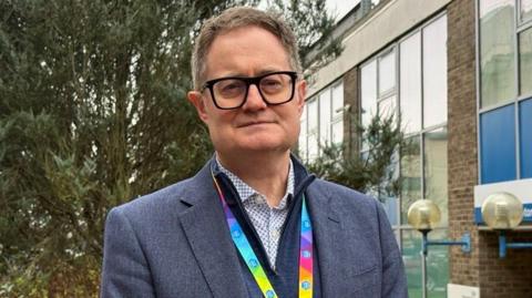 Paul Grundy poses for a photo outside Southampton General Hospital. He is not smiling and wears a dark jacket over a cardigan and patterned shirt, as well as a colourful lanyard.