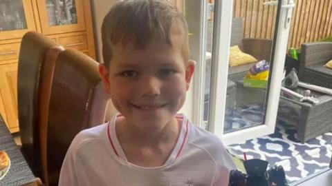 Eight year-old Joshua in a white shirt inside a house. He is smiling.