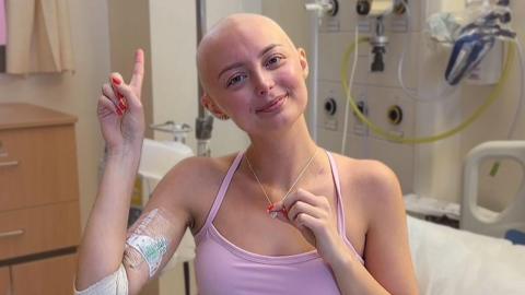 Person sitting in a hospital room wearing a pink top, with a bandaged arm and medical dressing visible. The person holds a necklace while raising one finger. Hospital equipment, tubing, and a bed are visible in the background.