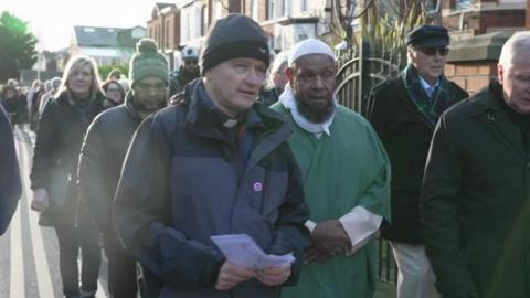 A large group of people walks along a footpath and on the edge of a road. At the front of the group is a