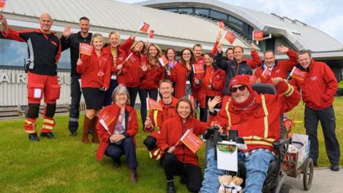 A group photo of 17 members of the charity and Steven Webb who is sitting in his wheelchair outside the headquarters. They are waving at the camera and smiling and some of them are waving small branded flags. Mr Webb has two small teddies tied to the front of his chair.