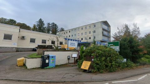 Cream buildings, with a large green bush in the foreground. A white sign, with blue writing reads 'Belford Hospital'.