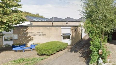 A single-storey, cream-coloured stone building, with a paved area to the front where there is a blue 'S'-shaped bench.