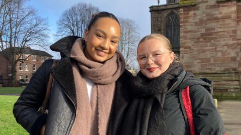 Two young women standing outside outside a cathedral. The woman on the left is wearing a black aviator jacket and a blush coloured scarf. The woman on the right is wearing circular, clear-framed glasses and a black coat, with a black scarf, she has a red bag over her shoulder. They both have their hair tied back and are smiling into the camera. The sky is blue in the background and there a couple of trees as well as a red brick building.