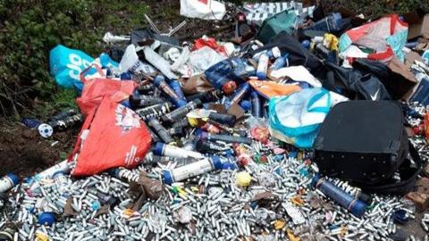 A large selection of rubbish on the ground, with lots of metal canisters. There are bags. Grass is all around it. 