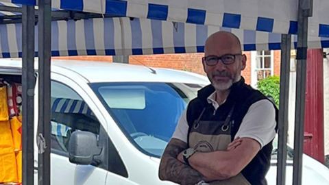 Robert Bird - a man with dark-framed glasses in a white t-shirt and dark gilet with an apron on - is standing behind a market stall. On the table are wood boards, mainly bread boards and chopping boards. Over him is a blue and white striped covering, sheltering the stalls. Behind him is a white van parked on a street