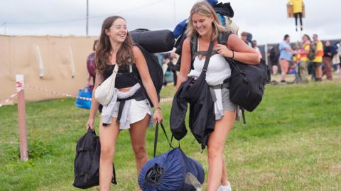 Two women carrying camping equipment walking through a field. There are people behind and red guide tape.