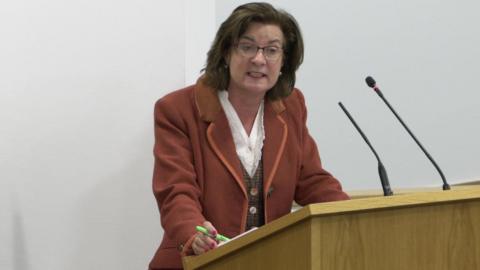 Eluned Morgan responding to Darren Millar during First Minister's Questions in the Senedd. She is standing at a light coloured wooden lectern with two microphones on it and holding a green pen in her right hand. 