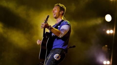 Chris Martin smiling and playing guitar wearing a blue top with black jeans with atmospheric lighting and mist behind him on stage