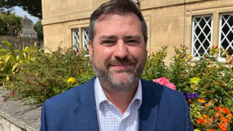 Kevin Guy. He has short dark hair and a beard. He is wearing a checked shirt and a navy blue jacket. He is looking at the camera and smiling.