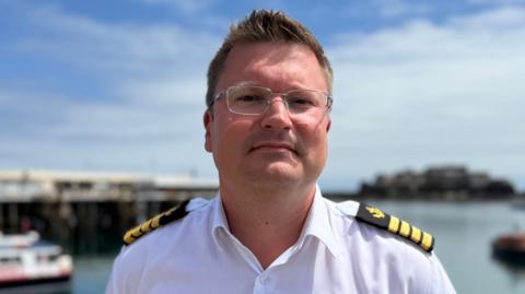 Head of Guernsey Coastguard James Way. He has short brown hair and silver glasses. He is wearing a white shirt with black and yellow epaulettes. the background of the image is blurred but parts of the harbour, like the water and boats are visible.