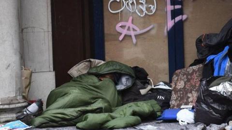 A rough sleeper is pictured lying on the ground in a green sleeping bag, surrounded by plastic bags, bin bags, a cushion. A pigeon is sitting on the sleeping bag.