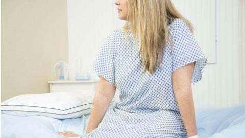 A woman wearing a white and blue hospital gown sits on the edge of a patient bed with her legs hanging down and her hands resting on the edge. She is looking to the left. The image is cut off above her nose. She has long, straight blonde hair.
