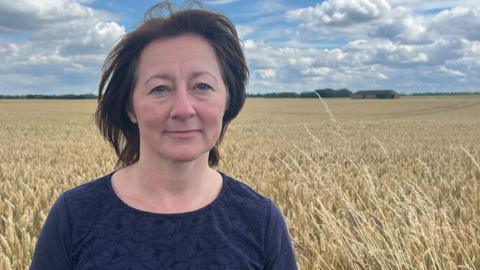 A woman with dark hair and wearing a blue t-shirt standing in front of a large field of golden wheat