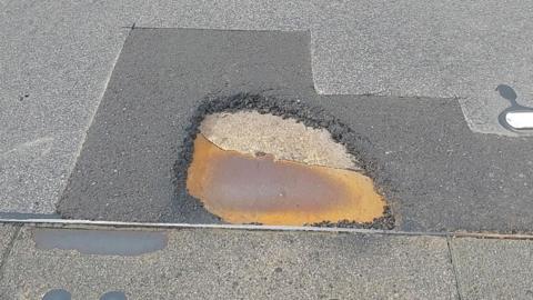 A large pothole in the tarmac with patches of repair work previously done around it