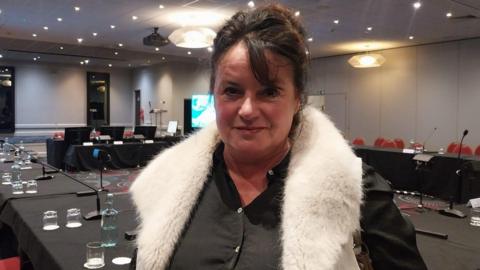 Brown haired woman looking at the camera with a soft smile. She is wearing a white furry gillet and a black shirt. She is standing in a meeting room, behind her are multiple tables, chairs and microphones. 