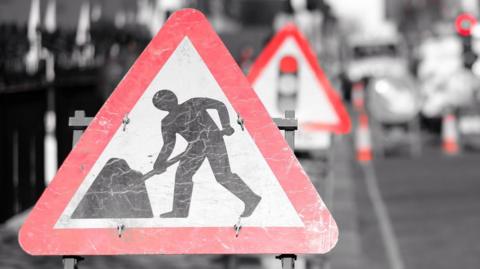 A triangle roadworks sign in the foreground, showing a figure of a man with a shovel in black in the middle and a red line around the triangle. Beyond is a blurred out road and other road signs. 