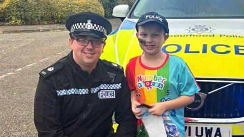 PC Brock is wearing his black and white police uniform. He is kneeling down in front of a police car and standing next to him is 7-year-old Luke. Luke is being treated for cancer and has a police cap on and a feeding tube on his face. 
