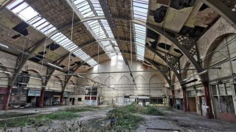 The inside of a disused indoor market building. Greenery is growing out of the grown and there is damage to the ceiling.