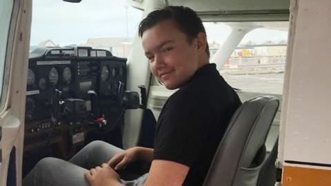 Chris Tromp is sitting in the cockpit of a small aeroplane with its door open. He is smiling and wearing a black t-shirt and jeans.