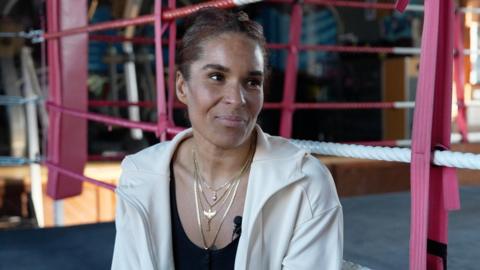 Lesley Sackey sits on the edge of a boxing ring, wearing a white jacket and layered necklaces, with pink boxing ropes in the foreground.