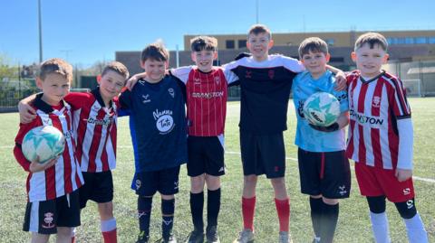 Seven boys lined up in a semi-circle on a football pitch. Two are holding blue footballs. Some are wearing a Lincoln City football kits. They are all smiling.