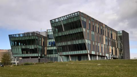 A large three-sectioned building, with brown panelling and glass frontage, with a blue BT sign in the middle. The fronts of the two end sections are sloped outwards so the top floor juts out further than the lower floors. There is a grass area in front of the building which leads down to the River Tyne, although that is out of shot. It is a cloudy but dry day.