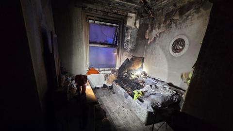 Bedroom with bed and dressing table and window at one end left charred after a fire