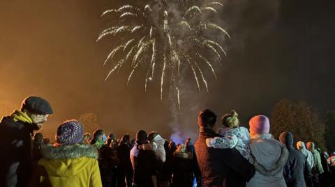 A group of people are gathered outdoors at night to watch a firework display. They are variously wearing coats and hats. There is a firework bursting in the air above the crowd.