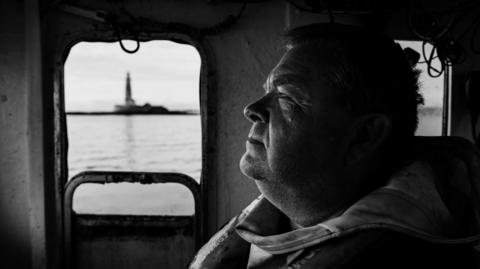A black and white photograph of a fisherman looking out to sea in the cabin of his fishing boat. A lighthouse can be seen through the window behind him. The sea looks calm. 