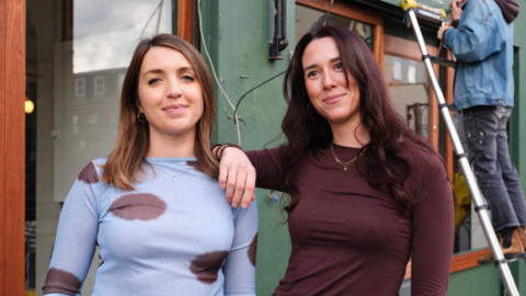 Two woman in front of a green shop, with someone on a ladder in the background. The woman on the left is wearing a blue shirt with brown dots, and the lady on the right is wearing a brown long-sleeve t-shirt. 