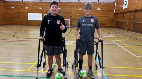 Two teenage boys with frame mobility aids stand next to each other in an indoor sports hall. They are both smiling and wearing football kits, and both have a green and white football at their feet. The boy on the left has dark hair and is giving a thumbs up, while the boy on the right has dark blond hair.