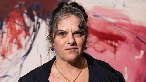 Dame Tracey Emin is seen wearing a black blazer over a dark top and a gold necklace. She stands in front of a large abstract painting with red and blue brushstrokes.