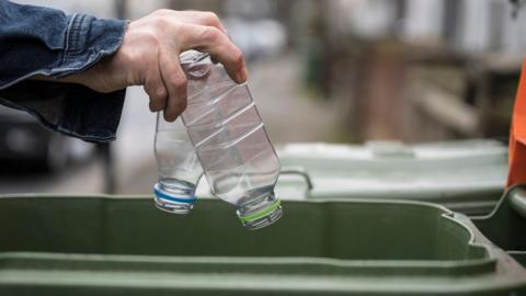 A hand dropping two plastic bottles into a green waste bin