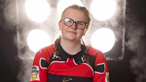 The picture shows a person wearing a red and black sports jersey with visible sponsor logos. The jersey has black and white detailing on the shoulders and sleeves. The individual has braided hair and is seated against a dramatic backdrop featuring six bright circular lights arranged in two vertical columns of three.