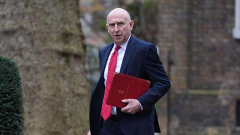 John Healey looks towards the camera as he carries a red folder