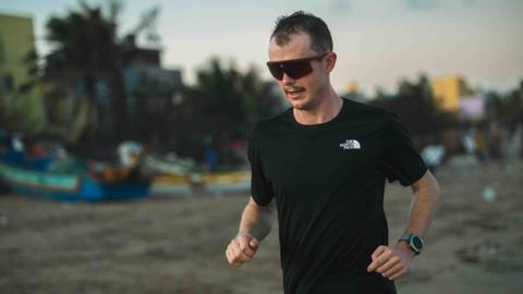 Will Robinson wearing a black North Face t-shirt, watch, and large black sunglasses. He is running along a beach at dusk. It is dimly lit, and in the out-of-focus background there are colourful boats resting on the sand and large palm trees.