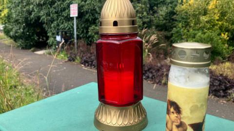 A close-up of two candles left at the scene of a man's murder in Fisher Park, Newry city.  One of the candles is in a red and gold glass lantern and the other is in a clear glass and gold jar and is decorated with a cherub-like figure.   A shrub-lined path in the park is partially visible behind the candles. 