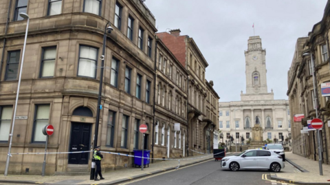 A street in Barnsley, with a police officer standing guard on the corner. A police cordon extends around a pavement on the left. A grand civic building is visible in the distance.