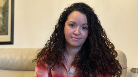 Margot Robert, with long brown curly hair and sitting on a sofa, looks at the camera