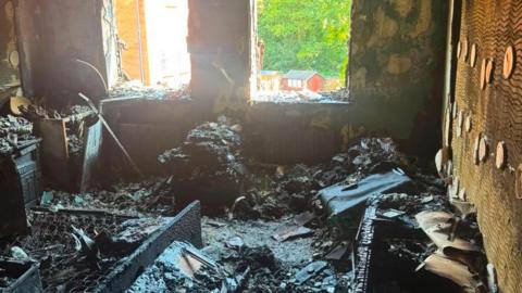 A fire-damaged bedroom, with charred remains. There are two windows, that have been destroyed. You can see through the windows to sheds and trees. 