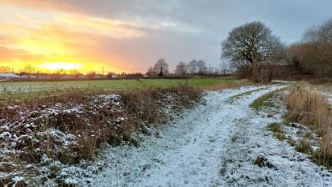 Snow lies on a country path as the sun sets with an orange and pink glow beyond a field to the left