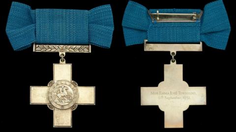 The front and back of the George Cross which is made of pale golden coloured metal mounted on a blue bow with a pin. On the front is an engraving showing a figure on a horse and on the reverse words are engraved reading 'Miss Emma José Townsend, 6th September, 1932.