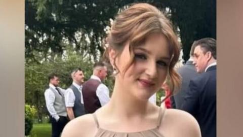Lainie is wearing a beige dress and holding a bunch of dried flowers. She smiling at the camera. Her brown hair is tied up with the two front pieces down and curled. It seems she is a bridesmaid at a wedding.