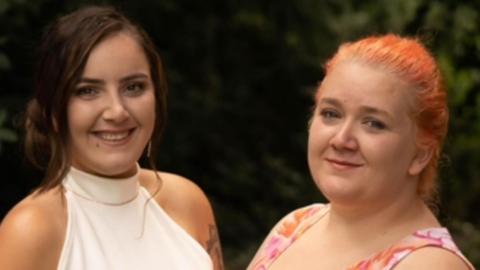 A close-up photo of Alex Whyte and Rhiannon Whyte, who are dressed up for a special occasion. Rhiannon, on the right, has orange-dyed hair tied back and a pink, white and orange sleeveless dress on. Alex, on the left, is wearing a white halter neck and has her brown high tied back. Both are slightly facing each other and smiling
